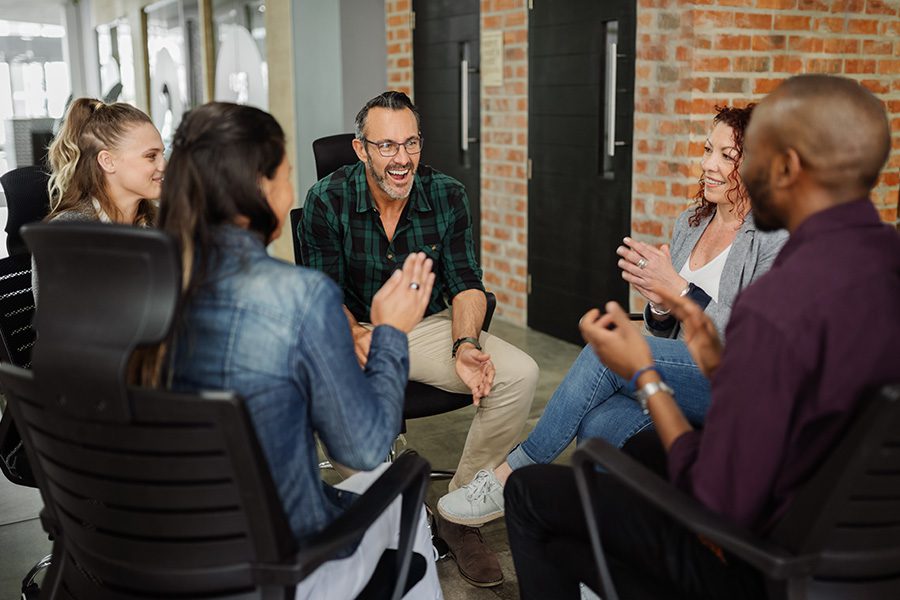 Employee Benefits - Group of Business People Sitting Together in a Circle in a Modern Office With Exposed Bricks Having a Casual Meeting