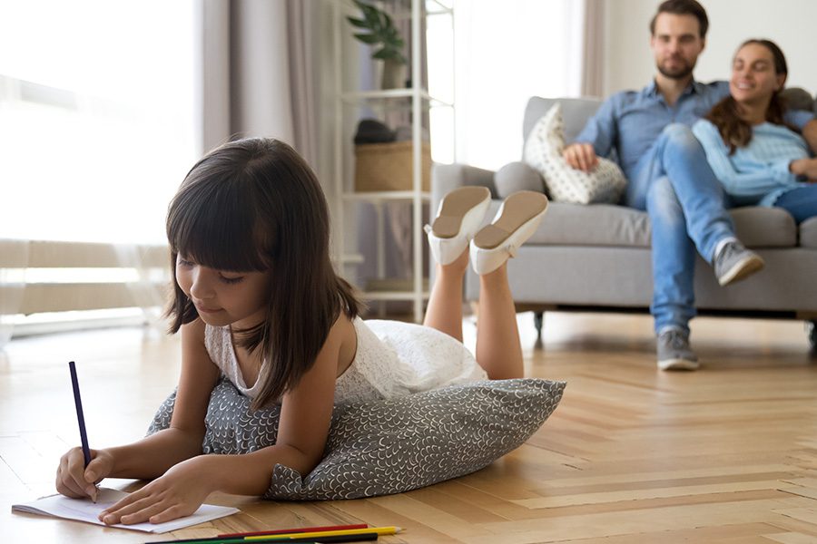 Personal Insurance - Father and Mother Sitting on a Sofa Together While Observing Their Daughter Draw Pictures on a Paper While Resting on a Pillow on the Floor